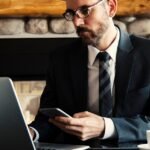 Businessman in formal attire working on a laptop and smartphone at an indoor office desk.
