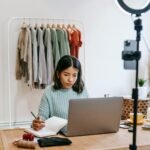 Focused young woman working on laptop in home office, surrounded by clothing rack and camera setup.